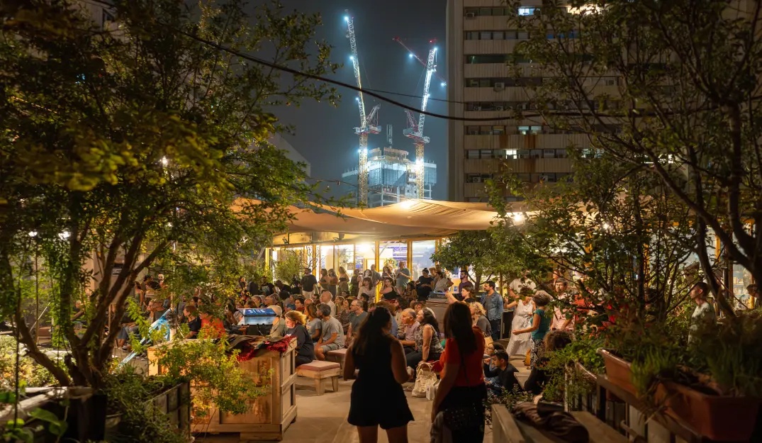A lively nighttime gathering in The Terrace of Muslala filled with people seated under canopy shades, surrounded by greenery and city buildings, with illuminated construction cranes visible in the background.