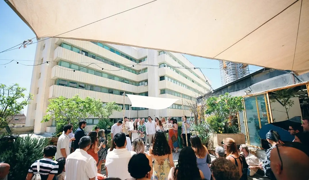 A wedding ceremony taking place at The Terrace of Muslala, with guests gathered under a large canopy and surrounded by greenery, against the backdrop of tall urban buildings.